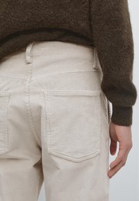 Pantalon en velours côtelé crème avec une surface texturée, comportant deux poches arrière, des coutures visibles et une taille ajustée.