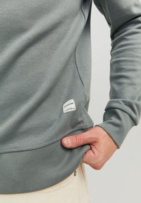 Homme portant un sweat-shirt gris avec une étiquette blanche Jack & Jones près de l'ourlet, main saisissant le bord inférieur sur un fond de pantalon beige.