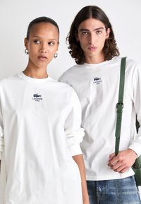 Baltos ilgos rankovės marškiniai su išsiuvinėtu Lacoste logotipu, su laisvu pjūviu ir sukabintomis rankovėmis. Vyrų modelis laiko žalią diržą krepšį.