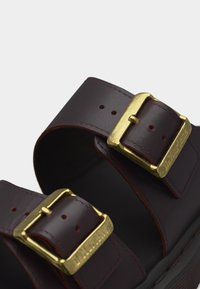 Λουριά από δέρμα σε σκούρο καφέ με δύο χρυσά μεταλλικά αγκράφες για ρυθμιζόμενη εφαρμογή, εμφανίζοντας λεπτή ραφή και τρύπες σε ανοιχτό φόντο.