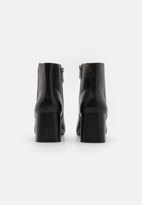 Botas de tobillo de cuero negro con un alto tacón bloque, con cremalleras laterales y textura suave. El diseño es elegante y moderno.
