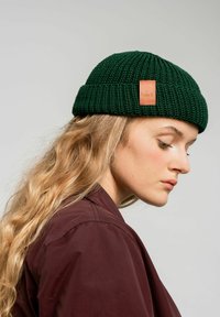 Zielona czapka dzianinowa z podwiniętym mankietem i skórzaną etykietą z boku. Gładka tekstura, okrągły kształt i średnia grubość.
