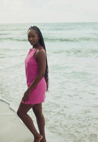 Rosa ärmlös klänning med avslappnad passform, står på en strand vid vattnet. Modellen har långt flätat hår och är barfota på blöt sand.
