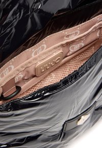 Interno di una borsa in vernice nera con una fodera a motivo floreale beige, dotata di un'etichetta brandizzata, di una tasca a rete e di una finitura lucida.
