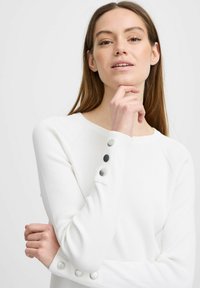 Pull en maille blanche à côtes avec de longues manches, doté de trois boutons gris sur chaque poignet. Texture douce avec une coupe décontractée et un col rond.