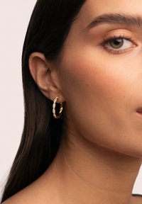 Pendiente de aro de oro con un diseño retorcido, con detalles de piedras preciosas claras. El pendiente cuelga de una oreja perforada, destacando su textura.