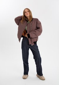 Lila bomberjacka med dragkedja fram, ribbade muddar och två sidofickor, matchad med mörka jeans och beiga sneakers.