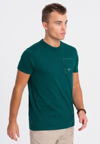 Tmavě modro-zelené tričko s krátkým rukávem vyrobené z bavlny, s kulatým výstřihem a kapsičkou na levé straně hrudi s textovým detailem. Hladká textura, přiléhavý střih.
