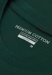 Zielona etykieta z premium bawełny na odzieży z białym tekstem informującym o "PREMIUM COTTON", "Intimissimi", "UOMO" oraz rozmiarze "M". Gładka tekstura.