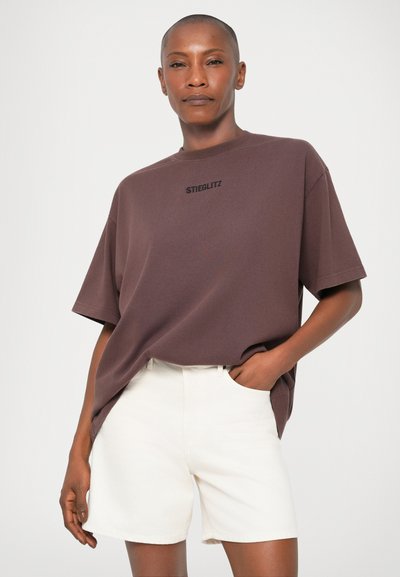 Camiseta oversized marrón con el logo "STIEGLITZ" en negro, combinada con pantalones cortos blancos. La tela parece suave; la camiseta tiene un escote redondo y mangas cortas.