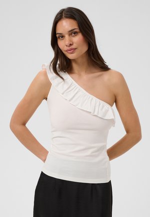 Femme portant un haut blanc à une épaule avec un détail volanté et un pantalon noir, debout les mains derrière le dos sur un fond clair.