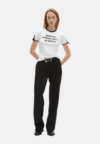 Hvid t-shirt med sort kant og tekst, der siger "IMOD AL AUTORITET BORTSET FRA KOOPLES," parret med højtaljede sorte bukser og sorte sko.
