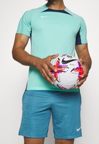 Ropa deportiva de Nike que incluye una camiseta verde menta con mangas texturizadas y detalles en azul marino, combinada con pantalones cortos azules, sosteniendo un balón de fútbol colorido.