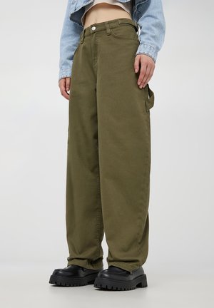 Pantaloni cargo larghi color verde oliva in tessuto di cotone, con tasche laterali e chiusura frontale con bottone, abbinati a scarpe platform nere chunky.