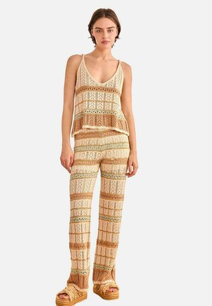 Mujer con top de tirantes de crochet a rayas beige y marrón y pantalones a juego, con sandalias de plataforma, sobre fondo blanco.