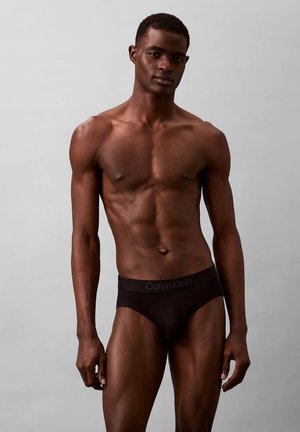 Slip di cotone nero con un elastico brandizzato, design aderente e stile minimalista. Il modello è in piedi contro uno sfondo neutro.