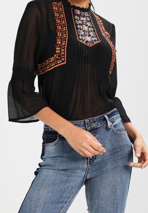 Blouse noire transparente avec broderie florale sur la poitrine et les épaules, manches trois-quarts évasées, associée à un jean bleu taille haute.