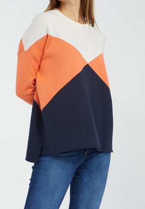 Nainen pukeutuneena väriblokkipuseroon, jossa valkoinen, oranssi ja tummansininen, yhdistettynä sinisiin farkkuihin, seisoo yksinkertaisen vaalean taustan edessä.