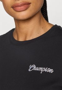 Crni pamučni majica s okruglim izrezom i bijelim izvezenim "Champion" logom na prsima. Glatka tekstura, ležeran kroj.
