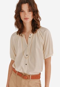Cremfarbige Bluse mit Knöpfen, gepufften kurzen Ärmeln und einem Rüschenausschnitt. Zugeordnet mit einem hellbraunen Gürtel, der eine goldene Schnalle im Seil-Design hat.