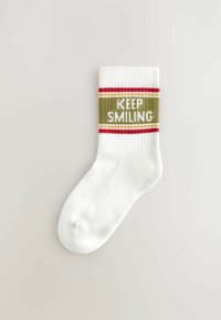 Biała bawełniana skarpetka z prążkowanym ściągaczem. Zawiera napis "KEEP SMILING" na białym tle, otoczony czerwonymi i żółtymi paskami.