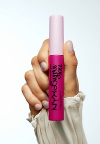 Bright pink läppstift i en matta förpackning, med svart varumärkesetikett och ett ljusrosa lock, hålls i en hand med mjuka, naturliga naglar.