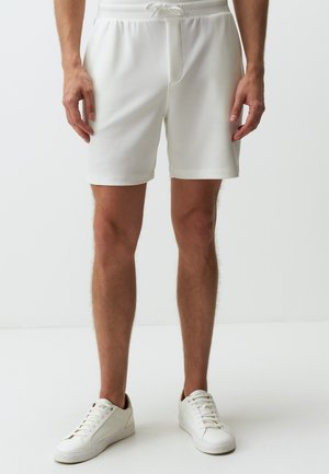 Shorts de sport blancs en tissu lisse, avec une taille élastique et un cordon de serrage, arrivant juste au-dessus des genoux. Portés avec des baskets blanches.