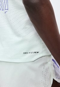 Gros plan d'une personne portant un vêtement de sport blanc perforé avec l'inscription "DRI-FIT ADV" près de l'ourlet et des détails graphiques bleus sur le short.
