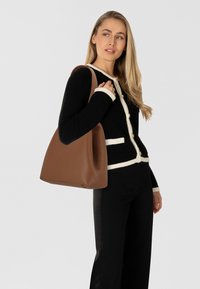 Borsa tote in pelle marrone con texture morbida, apertura ampia e forma curva. Indossata sulla spalla, abbinata a un cardigan nero con trama.