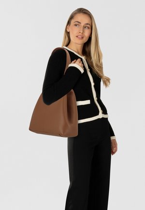 Borsa tote in pelle marrone con texture morbida, apertura ampia e forma curva. Indossata sulla spalla, abbinata a un cardigan nero con trama.