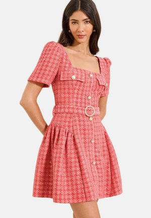 Žena v ružových textúrovaných krátkych rukávoch s perlovými gombíkmi, štvorcovým výstrihom, vreckami s klopou a zodpovedajúcim opaskom s perlovou prackou.