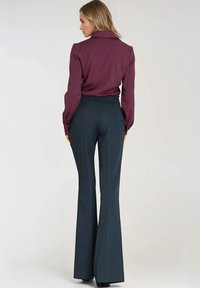 Kobieta stojąca tyłem, ubrana w bordową bluzkę z długim rękawem i ciemne, wysokie spodnie w kształcie dzwonów na jednolitym tle.