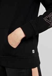 Sweatshirt noir avec poignets et ourlet côtelés, doté d'une poche avant et d'une petite étiquette blanche. Le design inclut un logo discret de couleur claire sur la manche.