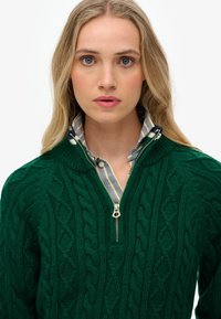 Groene kabelgebreide trui met een half-rits ontwerp, met daaronder een gestreept collshirt. Gevoerde textuur en metalen ritsdetails.