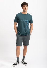 Teal katoenen t-shirt met een grafisch ontwerp in blauw en wit, gecombineerd met grijze cargoshorts, zwarte sneakers en grijze sokken.