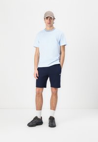 Tricou din bumbac de culoare albastru deschis, cu guler rotund, asortat cu pantaloni scurți bleumarin. Complementat cu adidași gri și o șapcă bej.