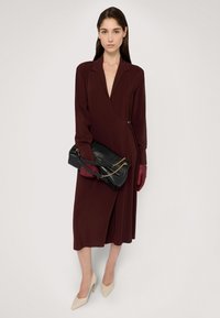 Bordowa długa sukienka z głębokim dekoltem w kształcie litery V, z paskiem w talii, w połączeniu z czarną torebką i czerwonymi rękawiczkami. Modelka w beżowych szpilkach.