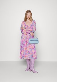 Vestido midi floral com padrões em roxo, rosa e amarelo; mangas longas e decote em bico. Combinado com uma mala estruturada em azul claro e botas brilhantes em lavanda.