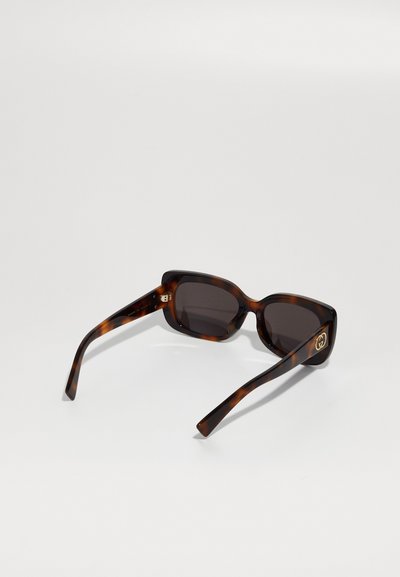 Lunettes de soleil en écaille de tortue avec un design carré, verres sombres et accent en logo doré sur la branche. Matériau plastique lisse et finition élégante.