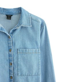 Lyseblå denimskjorte med krave, forlommer og knaplukning, der har et let tekstureret stof og synlige syningsdetaljer.