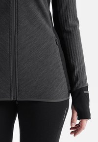 Giacca atletica a strati di colore grigio scuro con tessuto testurizzato, colletto alto, zip frontale e polsini con foro per il pollice. Presentate leggins neri aderenti.