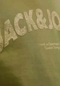 Tamsiai žalia medvilninė marškinėliai su tekstūruotais iškiliais užrašais "JACK&JONES" smėlio spalvos, o po jais mažesniu šriftu parašyta "Sukurta Kopenhagoje."