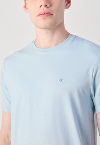 Hellblaues Baumwoll-T-Shirt mit rundem Halsausschnitt und kurzen Ärmeln. Verfügt über ein kleines gesticktes Logo auf der Brust. Weiche Textur.
