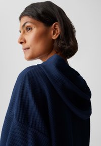 Donna con i capelli corti e scuri, che indossa un capo con cappuccio di un blu navy strutturato, che guarda verso sinistra contro uno sfondo chiaro e semplice.
