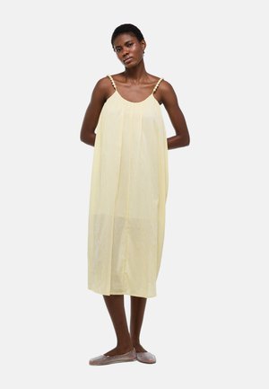 Femme portant une robe ample sans manches jaune clair avec bretelles perlées et chaussures plates tissées, debout les mains dans le dos.