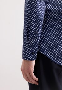 Mano di un uomo appoggiata sul fianco, indossando una camicia a maniche lunghe blu navy con pois azzurri e pantaloni scuri su uno sfondo neutro.