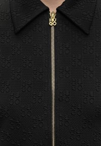 Κοντινή λήψη μαύρου υφασμάτινου σακακιού με υφή και χρυσό φερμουάρ με το όνομα της μάρκας "HUGO" σε κεφαλαία γράμματα.