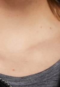 Šedá, texturovaná tkanina košile s kulatým výstřihem. Na jedné straně límce je viditelný zip s metalickými akcenty.