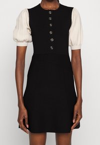 Robe sans manches noire avec six boutons en argent sur le devant, portée sur un haut crème à manches courtes et bouffantes.