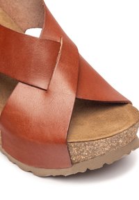 Brown leather wedge sandal featuring crisscross straps, a textured cork sole, and a rounded toe design. Smooth finish and sturdy construction.